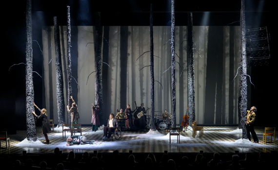 Skådespelare bland höga trädstammar på scen med snö, en kvinna ligger på golvet, band spelar i bakgrunden.