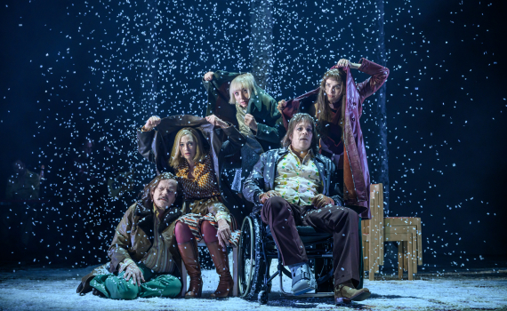 Fem personer, varav en i rullstol, skyddar sig mot snöfall med upplyfta rockar, scen med mörk bakgrund.