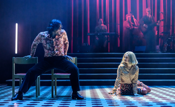 Man i mönstrad skjorta sitter gränsle över stol, kvinna i färgglad klädsel sitter på golvet, band i bakgrunden.