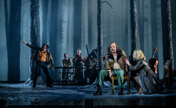 Man sitter på stol i skogsscen, kvinna hukar vid hans sida, musiker syns bakom bland trädstammar.