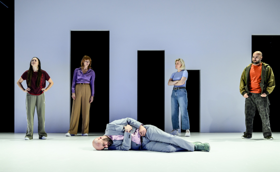 Fem personer står på scen, en man i kostym ligger ner medan de andra tittar åt olika håll.