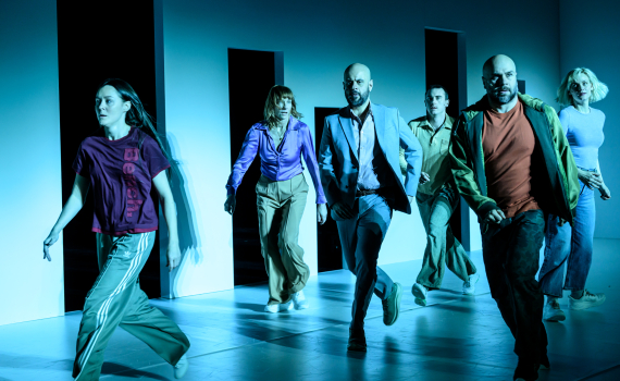 Sex personer springer mot kameran på en scen med blå belysning och svarta öppningar i väggen bakom dem.