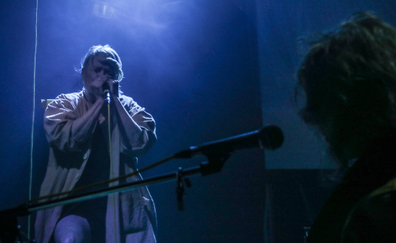 En kvinna som sjunger i ett blått upplyst rum, i förgrunden ser man siluetten av en musiker