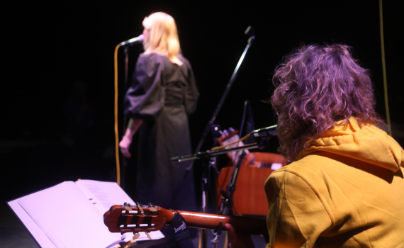 Ryggen på en musiker som spelar gitarr, i bakgrunden ser man en person som står vid en mikrofon 