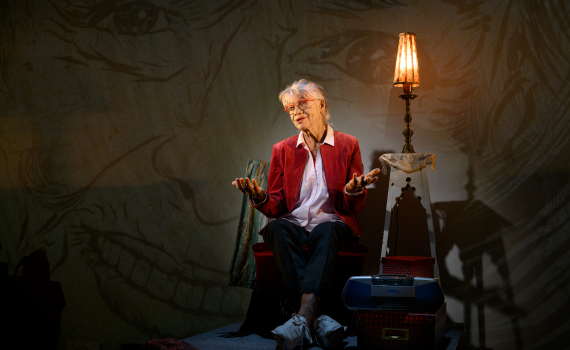 Iwa Boman sitter i en fåtölj, i bakgrunden anar man porträtt projicerade på väggen.