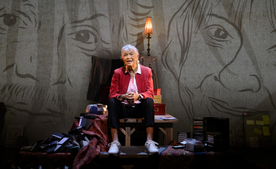 Iwa Boman sitter i en fåtölj, i bakgrunden ser man ritade porträtt på väggen.