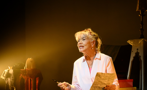 Iwa Boman sitter lutad mot en ställning hållandes ett papper. En varmt gult ljus lyser mot henne.