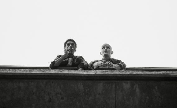 Två unga män lutar sig över ett räcke, en röker cigarett, den andra är skallig och tittar uppåt.