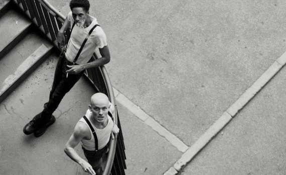 Två unga män i hängslen och t-shirts står i en trappa, båda röker och tittar uppåt.