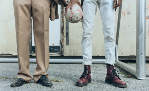 Två personer står bredvid varandra; en i beiga byxor och svarta skor, den andra i vita byxor och röda kängor, håller en sliten fotboll.