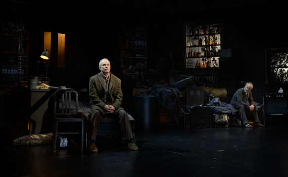 Två äldre män i scenmiljö med sängar och flaskhyllor; en sitter i kavaj, den andra i mörk tröja.