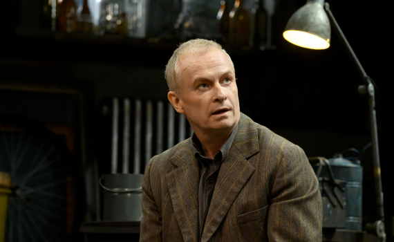 Medelålders man i brun kostym sitter på en säng och håller ett föremål i händerna.