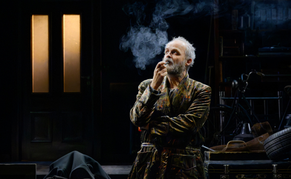 Äldre man med vitt hår och skägg står rökande i rock, omgiven av rekvisita och dimma.