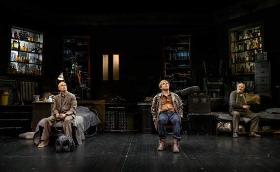 Tre män sitter utspridda på scen med flaskhyllor bakom; alla tittar uppåt.