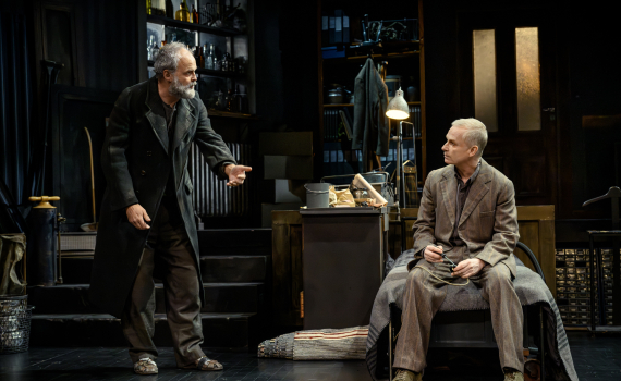 En äldre man i mörk rock gestikulerar mot en sittande man i brun kostym på en scen.