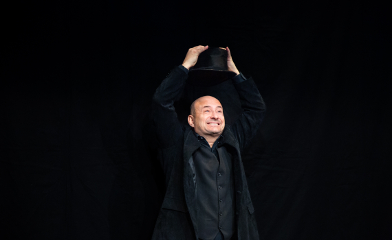 Alt Text Generator sade:  Skallig man i svart klädsel håller en hög hatt över huvudet mot svart bakgrund.