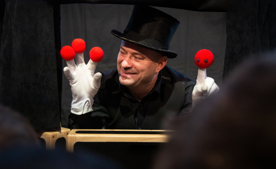 Man med hög hatt visar tre röda bollar på ena handen och en docka på den andra.