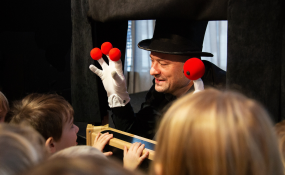 Man med hög hatt och vita handskar uppträder med röda bollar inför barn.