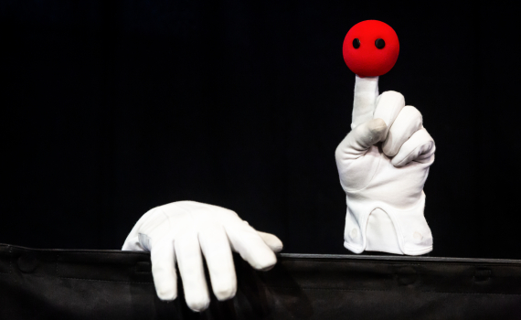 Vit handske håller upp en röd docka med svarta ögon framför svart bakgrund.