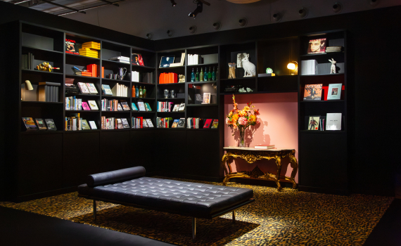 Bibliotekshörna med svarta bokhyllor, böcker, leopardmönstrad matta, svart schäslong och guldbord med blommor.