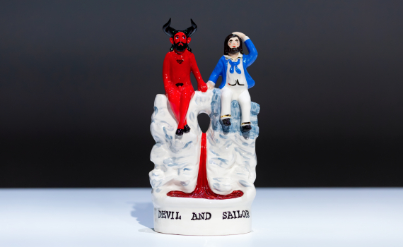 Glaserad figur av röd djävul och sjöman i blå jacka, sittande tillsammans, text ”Devil and Sailor”.