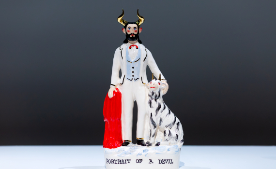 Glaserad figur av manlig djävul i vit kostym med horn, röd mantel och vit hund, text ”Portrait of a Devil”.