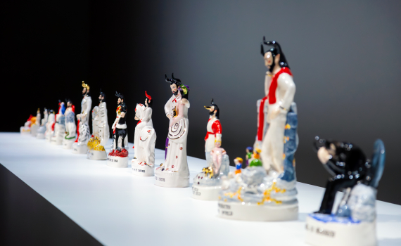 Rad av små glaserade djävulsfigurer i olika kläder och poser uppställda på vit hylla.