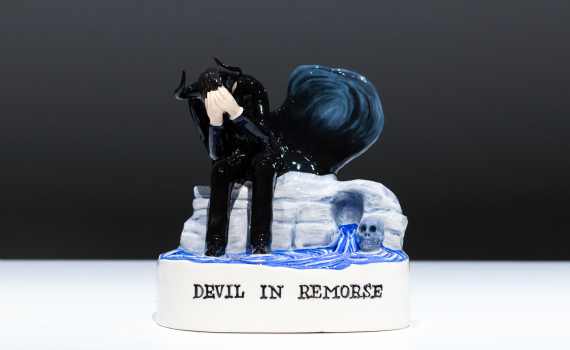 Glaserad figur av svartklädd djävul med horn, sittande med ansiktet i händerna, text ”Devil in Remorse”.