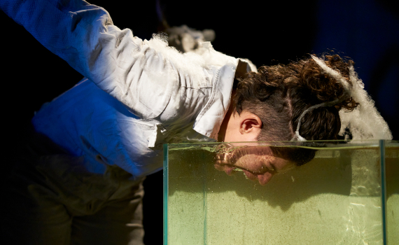 Person i vit overall lutar sig framåt med ansiktet nedsänkt i glasbehållare fylld med grumligt vatten.