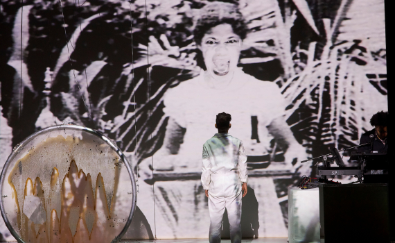 Person i vit overall står med ryggen mot kameran framför stor projicerad bild; musikutrustning och trumma på scenen.