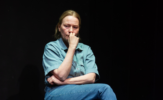 Medelålders kvinna i blå skjorta och jeans sitter på stol mot svart bakgrund, handen mot munnen.