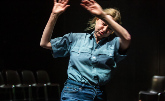 Medelålders kvinna i blå skjorta och jeans rör sig med armarna upplyfta på mörk scen.