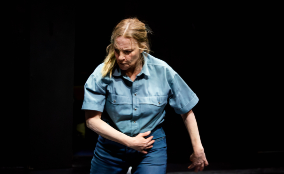 Medelålders kvinna i blå skjorta och jeans lutar sig fram med handen mot magen.