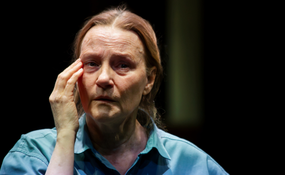 Medelålders kvinna i blå skjorta rör vid kinden med handen, mörk bakgrund.