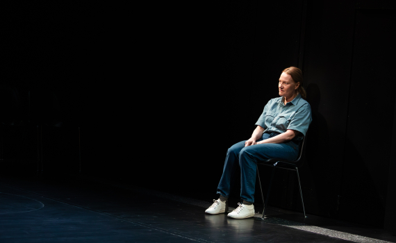 Medelålders kvinna i blå skjorta och jeans sitter ensam på stol i mörkt rum.