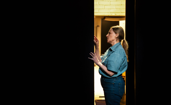 Medelålders kvinna i blå skjorta och jeans står i dörröppning med upplyst rum bakom.