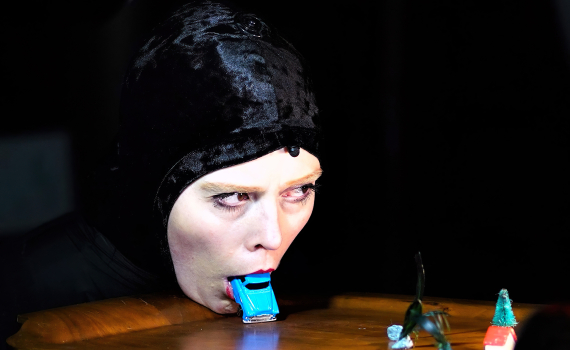 Kvinna i svart huvudbonad ligger med ansiktet mot ett bord och håller en blå leksaksbil i munnen.