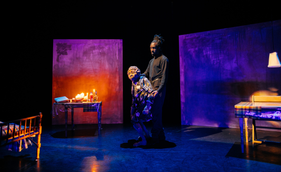 Man i svarta kläder står bakom en docklik äldre kvinna i blommig klänning på scen med säng och bord.