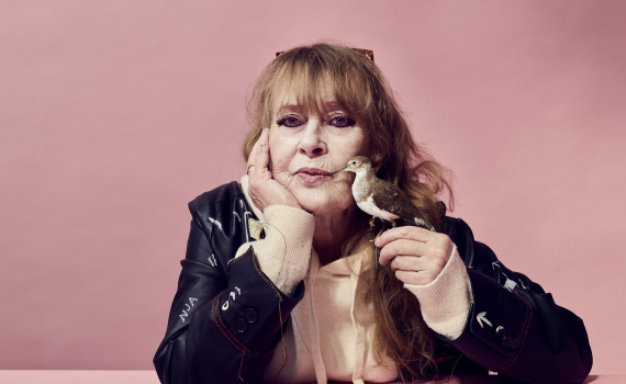 Kvinna i svart kavaj lutar huvudet mot handen vid ett rosa bord. Hon håller en liten fågel nära ansiktet mot en rosa bakgrund.