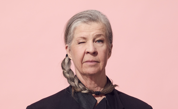 Person i svart kavaj med grå fläta sitter rakt fram och blinkar. Händerna är knäppta framför kroppen mot rosa bakgrund.