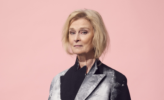 Kvinna i svart och vitmålad kavaj står i halvprofil. Minimalistiskt porträtt mot rosa bakgrund.