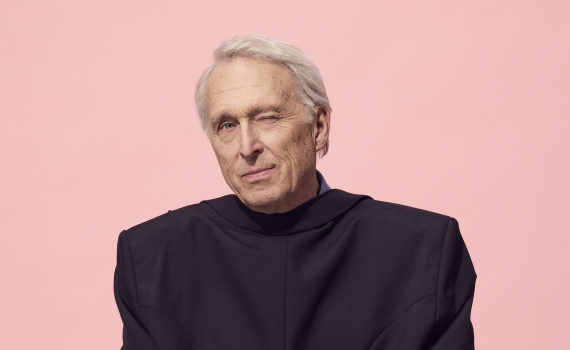 Man i svart kavaj sitter på en låda och blinkar mot kameran. Enkel studiobild mot rosa bakgrund.