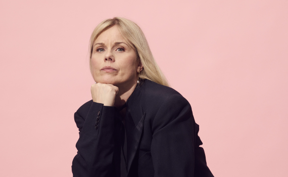 Kvinna i svart kostym sitter hukad på en scenlåda med handen under hakan. Porträtt mot rosa bakgrund.