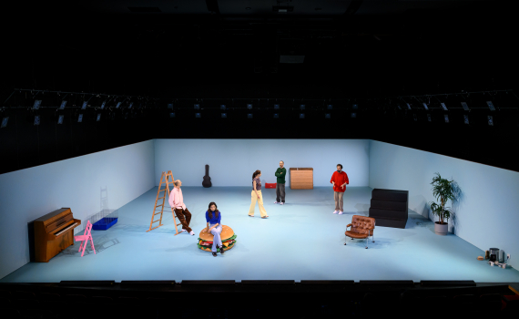 Helbild av scenrum med fem personer utspridda bland stege, piano, fåtölj och andra objekt.
