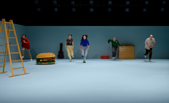 Fem personer springer över en scen med stege, jättelik hamburgare och rekvisita i en minimalistisk teatermiljö.