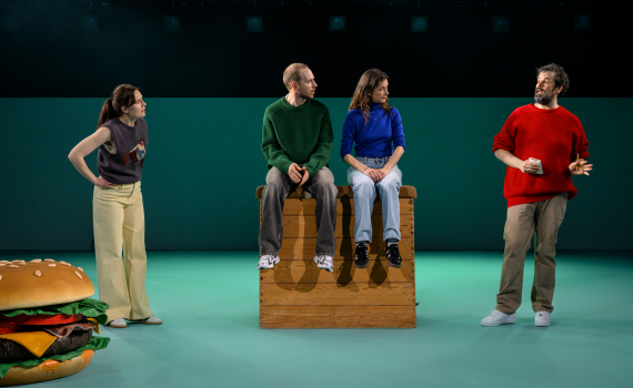 Fyra personer samtalar; två sitter på låda, en står bredvid och en gestikulerar i en öppen scenbild.