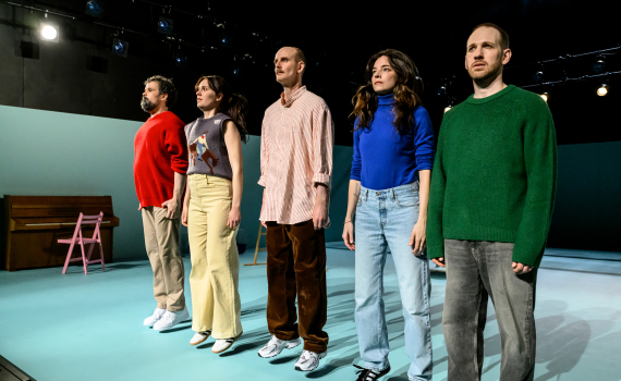 Fem personer står i rad på scen och blickar framåt, upplysta av starka scenlampor.