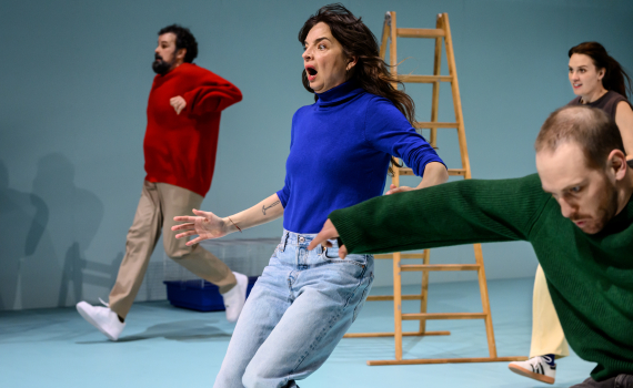 Tre personer i rörelse på scen; kvinna i blå tröja i fokus, andra springer förbi i bakgrunden.