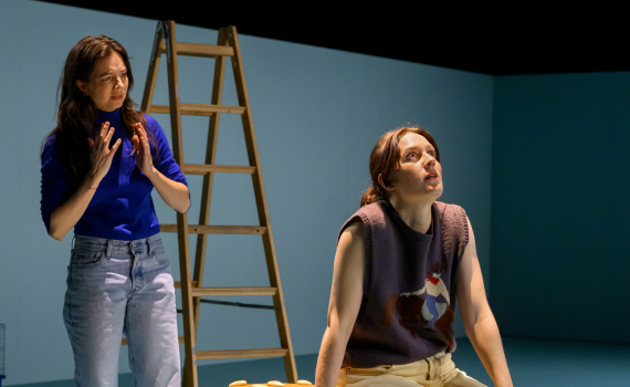 Två kvinnor vid stor hamburgarrekvisita; en sitter och ser upp, den andra står bakom med händerna lyfta.