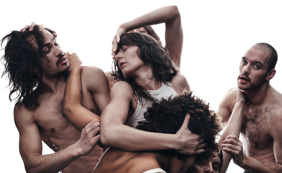 Fem vuxna dansare, män och kvinnor ca 20–35, i kroppsnära kläder rör sig tätt ihop med armar runt varandras huvuden och axlar.